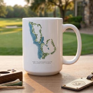 May include: White ceramic mug featuring a golf course map design in blue and green. The mug has a large handle and the text "THE PLANTERRA CLUB PEACHTREE CITY, GEORGIA". Golf tees and a leather tag are also visible.