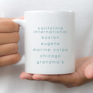 May include: White ceramic mug with the text "california international boston eugene marine corps chicago grandma's" printed in teal blue.