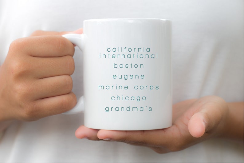 May include: White ceramic mug with the text "california international boston eugene marine corps chicago grandma's" printed in teal blue.