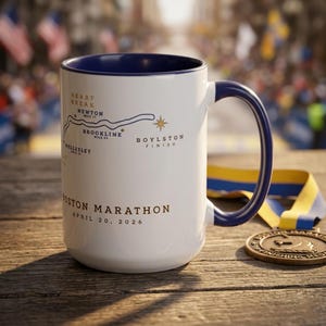 May include: White ceramic mug with a blue interior and handle, featuring a map of the Boston Marathon route. The mug includes text indicating the race date, April 20, 2026, and a gold medal and ribbon are visible.