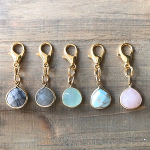 May include: Five gold keychains with different colored gemstone pendants. The pendants are teardrop shaped and include grey, light blue, white, and pink.