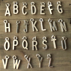 Puede incluir: Un conjunto de 26 abalorios de letras plateadas, cada letra del alfabeto está representada. Los abalorios están sobre una superficie de madera.