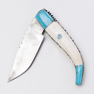 Op de afbeelding: Een vouwmes met een wit beenhandvat en een turquoise inleg. Het lemmet is gemaakt van damaststaal met een golvend patroon.