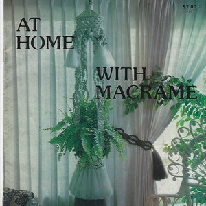 May include: A book cover with the title "At Home With Macrame" and a photo of a macrame plant hanger with a green plant hanging from it. The book is a "Classic Book" and is part of the "Great Home Projects" series.