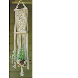 May include: A white macrame plant hanger with a wooden dowel and a green plant in a brown pot.