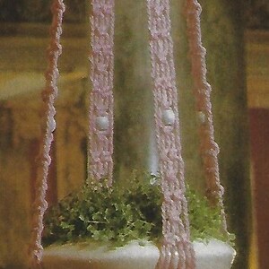 May include: A pink crocheted hanging planter with a white tassel. The planter is filled with green plants and has a white ceramic base.
