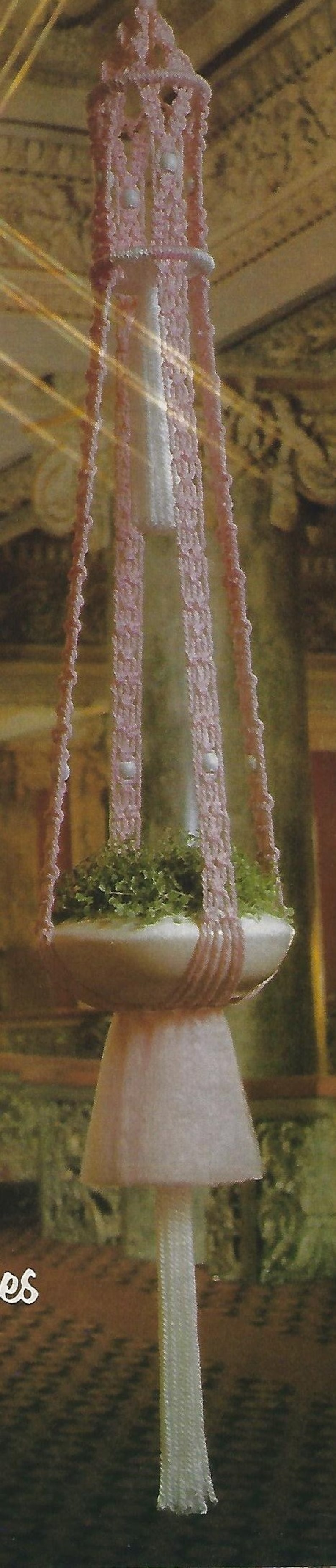 May include: A pink crocheted hanging planter with a white tassel. The planter is filled with green plants and has a white ceramic base.