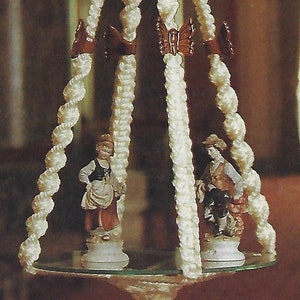 May include: A white macrame plant hanger with a round glass top and three legs. The hanger is decorated with small brown beads and two figurines. The hanger is hanging from the ceiling.
