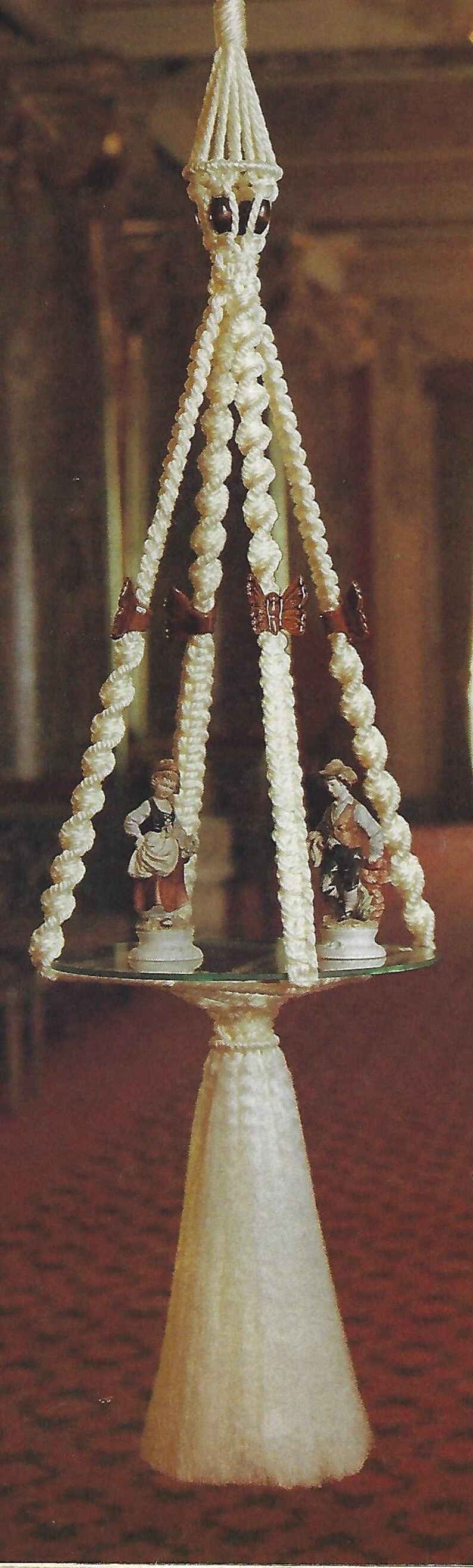 May include: A white macrame plant hanger with a round glass top and three legs. The hanger is decorated with small brown beads and two figurines. The hanger is hanging from the ceiling.