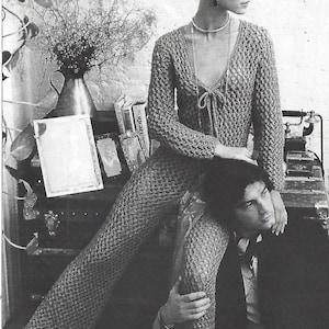 May include: A black and white photograph of a woman wearing a knitted jumpsuit. The jumpsuit is loose-fitting and has a V-neck. The woman is sitting on a man's lap. The photo has a vintage feel.