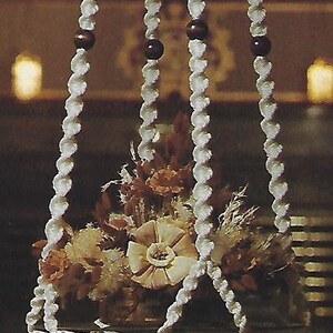 May include: A white macrame hanging planter with a white tassel. The planter is filled with dried flowers and is hanging from a ceiling.
