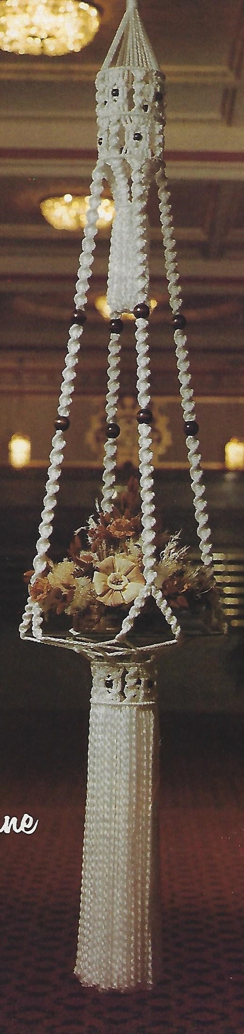 May include: A white macrame hanging planter with a white tassel. The planter is filled with dried flowers and is hanging from a ceiling.