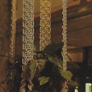 May include: A macrame plant hanger with a white and cream color scheme. The hanger is made of knotted cords and has a round pot at the bottom. The hanger is hanging from the ceiling and there are green plants growing in the pot.