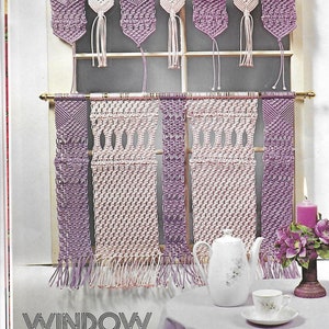 May include: A set of six macrame window hangings in shades of purple and pink. The hangings are made with a knotting technique and feature tassels at the bottom. The hangings are displayed on a window with a white frame. The text "WINDOW WHAT-KNOT" is printed below the hangings.