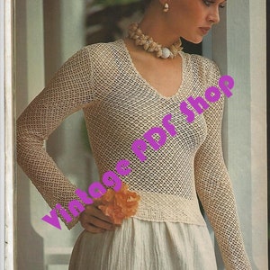 May include: A white crochet top with a v-neck and long sleeves. The top is made of a delicate, open-weave fabric. The model is wearing a white skirt and a shell necklace.