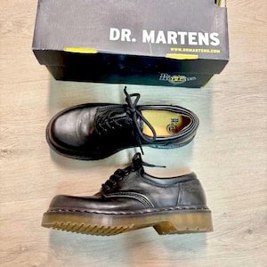 Peut inclure: Chaussures Dr. Martens en cuir noir avec un design à lacets. Les chaussures sont présentées avec leur boîte d'origine, qui affiche le nom de la marque. Les semelles sont de couleur marron clair.