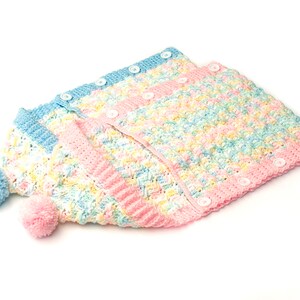 May include: A crocheted baby cardigan and hat set in a pastel rainbow color scheme with white buttons. The cardigan has a button-up front and the hat has a pom-pom on top.