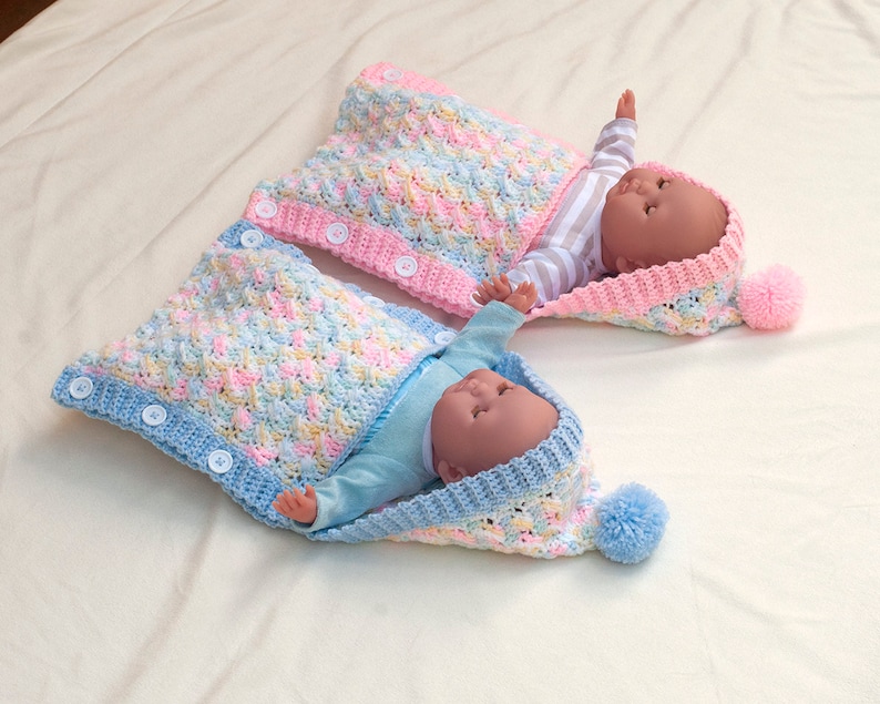 May include: Two crocheted baby sleeping bags with hoods and pom-poms. One is blue with white buttons and the other is pink with white buttons.