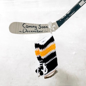 May include: A hockey stick with a white sign attached that says "Coming Soon ~ December ~". A black and white crocheted sock with yellow stripes hangs from the stick.