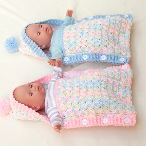 May include: Two crocheted baby sleeping bags with hoods and pom-poms. One is pink and the other is blue. Both have a striped pattern and are made with a textured stitch.
