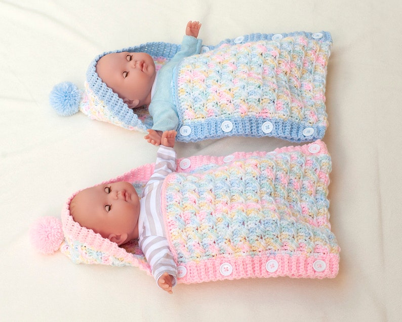 May include: Two crocheted baby sleeping bags with hoods and pom-poms. One is pink and the other is blue. Both have a striped pattern and are made with a textured stitch.