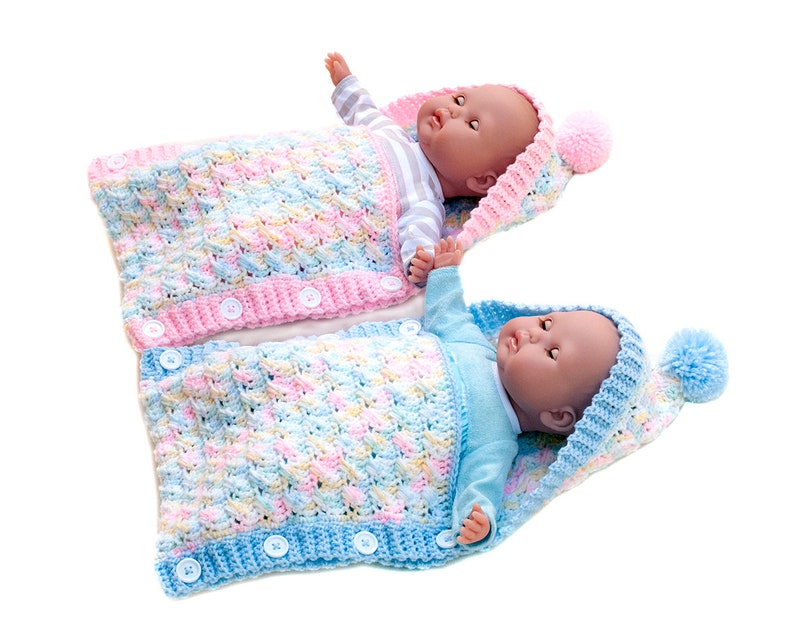 May include: Two crocheted baby sleeping bags with hoods and pom-poms. One is pink with yellow and blue accents, the other is blue with pink and yellow accents.