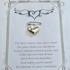 May include: A silver heart-shaped charm with the word "Sister" on it, attached to a safety pin. The charm is on a card with a heart and wing design, and a poem about remembrance.