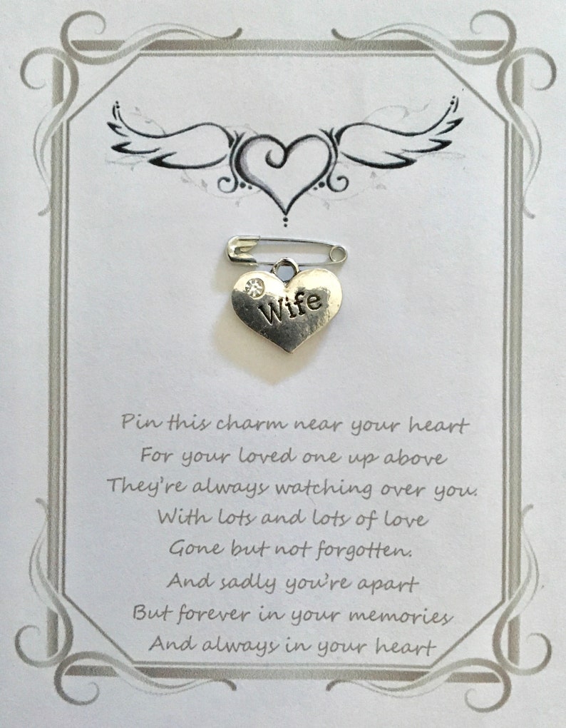 May include: A silver heart-shaped charm with the word "Wife" and a small crystal, attached to a safety pin. The charm is on a white card with a decorative border, a heart and wing design, and sentimental text.