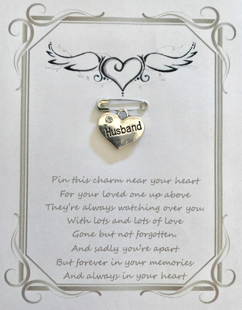 May include: A memorial pin with a silver heart charm that reads "Husband" attached to a safety pin. The pin is on a white card with a decorative border, a heart and wing design, and a poem.