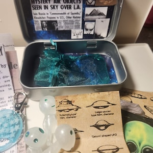 May include: A set of UFO themed items including a small tin box with a green mesh bag inside, three white alien figurines, a blue and white resin charm, and a card with illustrations of different UFO shapes and sizes. The card includes text that reads "Adamski type 10m. diameter", "Large saucer-type 50m. diameter", and "Saturn'-shaped UFO".
