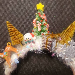 May include: A white headband with a Christmas theme. It features a small green Christmas tree with candy canes, a snowman, a reindeer, a snowflake, and gold glitter.