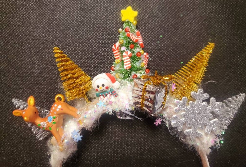May include: A white headband with a Christmas theme. It features a small green Christmas tree with candy canes, a snowman, a reindeer, a snowflake, and gold glitter.