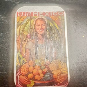 May include: A silver tin with a colorful image of a woman holding a bowl of fruit. The text "Visit Mexico" is printed on the tin.