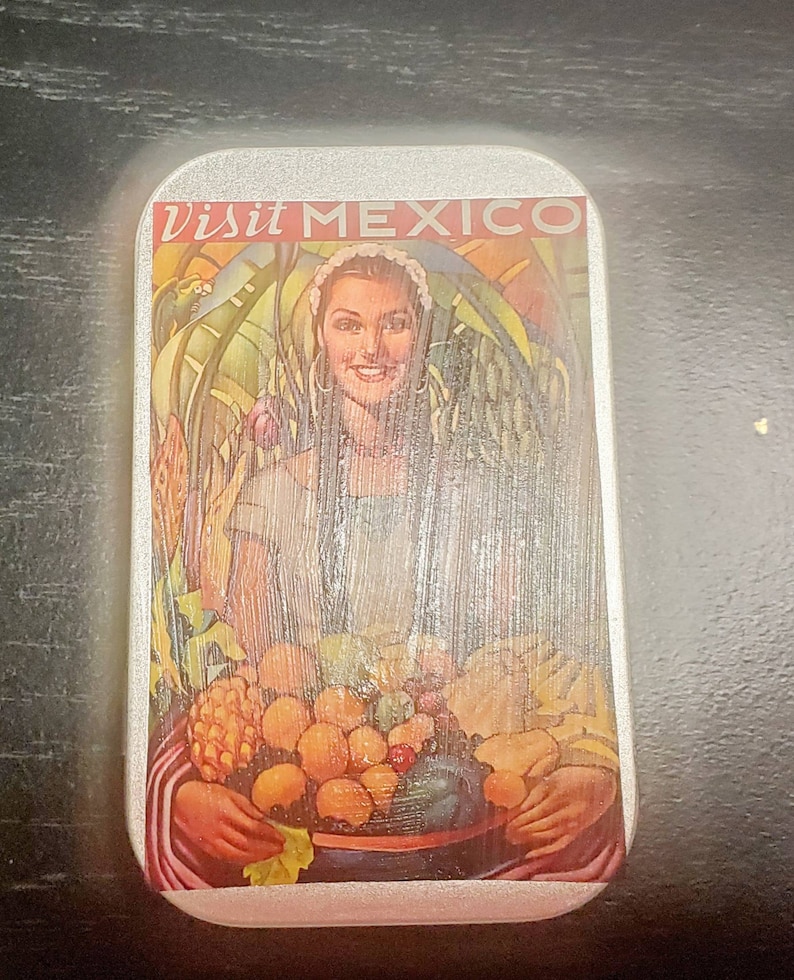 May include: A silver tin with a colorful image of a woman holding a bowl of fruit. The text "Visit Mexico" is printed on the tin.