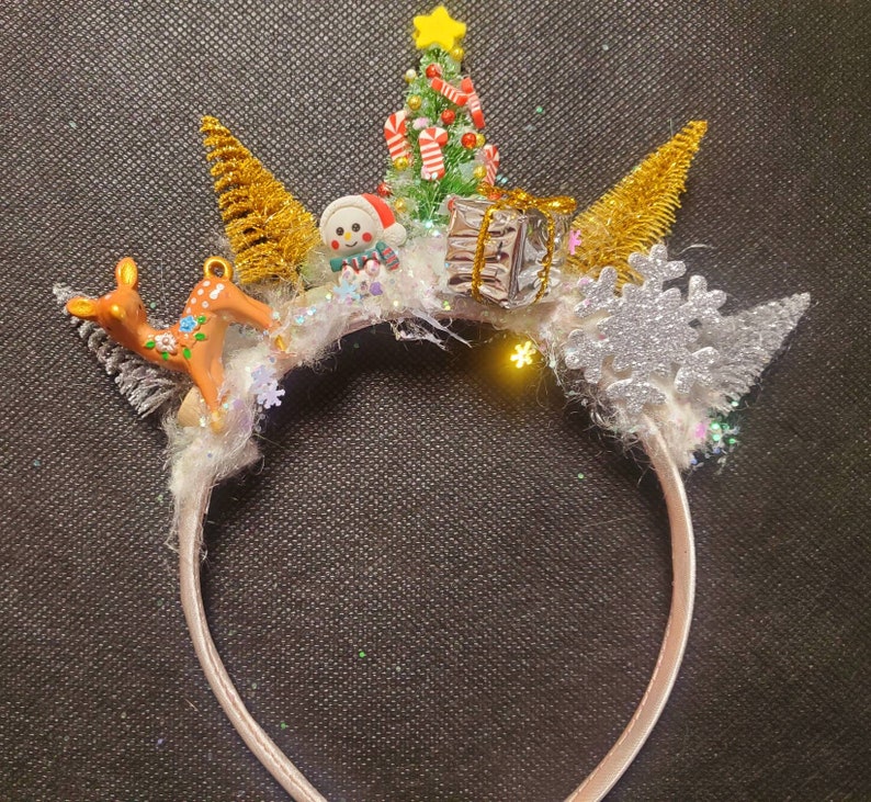May include: A white headband with a winter holiday theme. It features a small snowman, a Christmas tree, a reindeer, and a snowflake. The headband is decorated with glitter and faux fur.