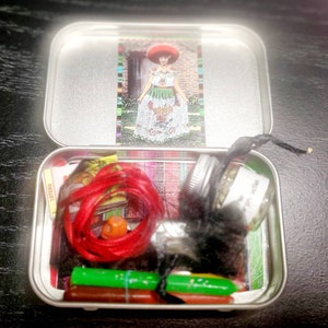 May include: A silver tin box filled with various items, including red string, green and orange crayons, a small glass jar with a black lid, and a black ribbon. The lid of the tin box features a colorful image of a woman wearing a traditional Mexican dress.