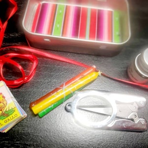 May include: A red ribbon, a small tin box with a colorful striped pattern, a small bottle with a silver lid, a pair of small silver scissors, and a small card with a rooster illustration and the text "EL GALLO" on it. The card is part of a deck of cards called "LOTERIA".