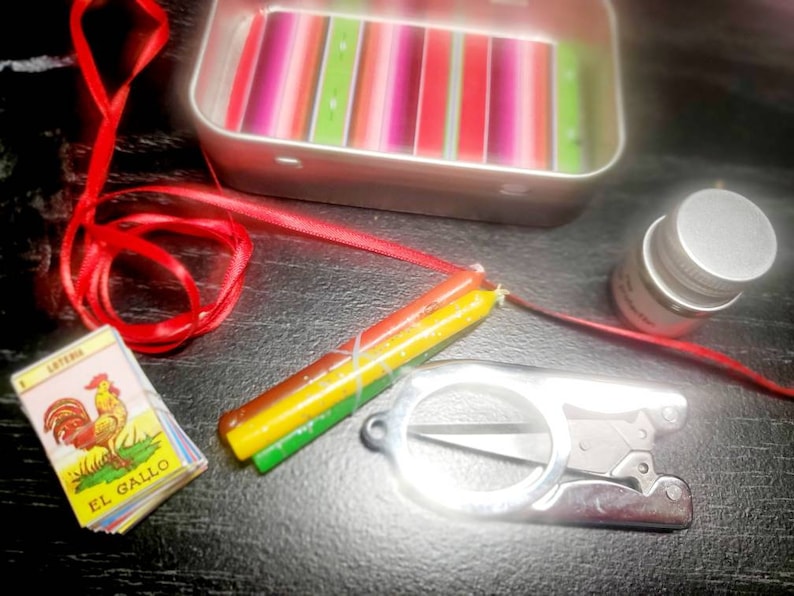 May include: A red ribbon, a small tin box with a colorful striped pattern, a small bottle with a silver lid, a pair of small silver scissors, and a small card with a rooster illustration and the text "EL GALLO" on it. The card is part of a deck of cards called "LOTERIA".