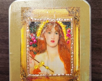 May include: A rectangular, gold-toned tin featuring a framed portrait of a woman with red hair and a yellow floral crown. The portrait is surrounded by a decorative gold frame with ornate detailing. The woman holds a sprig with yellow accents.