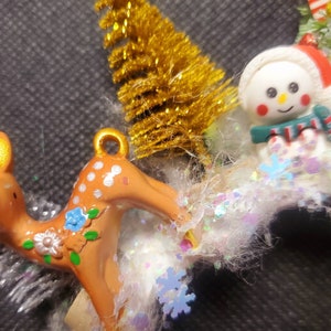May include: A Christmas ornament featuring a brown and white spotted deer with floral accents, a snowman with a red hat and a green scarf, and a gold glitter Christmas tree.