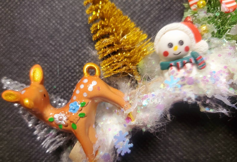 May include: A Christmas ornament featuring a brown and white spotted deer with floral accents, a snowman with a red hat and a green scarf, and a gold glitter Christmas tree.