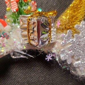 May include: A white headband with a snowman, a small Christmas tree, a silver gift box with a gold bow, and a silver snowflake.
