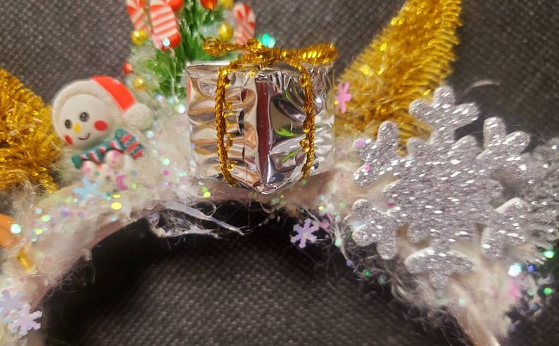 May include: A white headband with a snowman, a small Christmas tree, a silver gift box with a gold bow, and a silver snowflake.