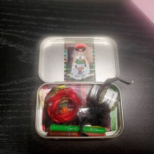 May include: A small, round, silver tin with a red, white, and green patterned lid. The tin is open and contains a red string, a small glass jar with a white label, and several green and red items.
