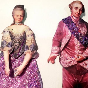 May include: Paper cutouts of a woman and a man in historical attire. The woman wears a gown with a purple glitter skirt and a bodice with floral accents. The man is in a pink suit with a purple glitter sash. Both figures have detailed facial features.