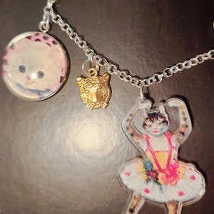 May include: A silver chain necklace with three charms: a round charm with a white kitten, a gold tiger head charm, and a clear charm with a ballerina cat wearing a pink and yellow tutu.