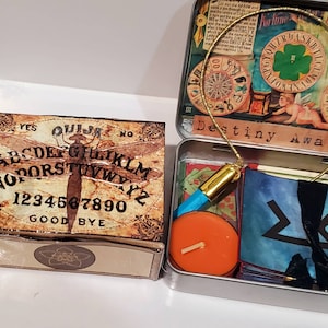 May include: A vintage-style wooden box with a Ouija board design featuring a dragonfly and the words "YES QUIJA NO" and "GOOD BYE". The box is open and reveals a smaller tin box with a gold chain, a teal blue bottle, a small orange candle, and a set of cards with a black and white symbol.