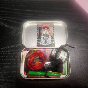 May include: A small, round, silver tin with a red, white, and green patterned lid. The tin is open and contains a red string, a small glass jar with a white label, and several green and red items.