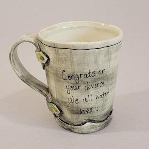 May include: A handmade ceramic mug with a gray and white textured finish. The mug features a handle and is decorated with green accents. The text "Congrats on your divorce, We all hated her!" is written on the side in black.