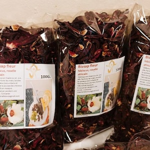 Peut inclure: Trois sachets transparents remplis de fleurs d'hibiscus séchées, rouge foncé. Chaque sachet porte une étiquette avec le texte "Bissap fleur hibiscus, roselle africain" et une image d'une fleur blanche et d'une boisson. Chaque sachet contient 100g de produit.